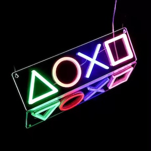Op de afbeelding: Een neon-bord met de PlayStation-knopsymbolen in verschillende kleuren. De symbolen zijn een driehoek, een cirkel, een X en een vierkant. Het bord hangt aan het plafond en is verlicht.