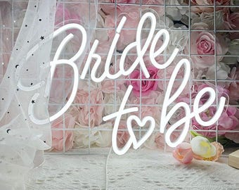 May include: A white neon sign that reads "Bride to be" with a heart symbol. The sign is mounted on a white grid against a backdrop of pink and white roses. A white veil with stars is draped on the left.