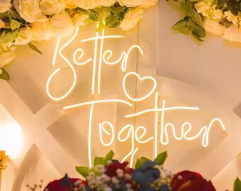 May include: A neon sign with the words "Better Together" in a cursive font, with a heart symbol. The sign is illuminated in a warm white color, set against a backdrop of white and cream-colored flowers and greenery.