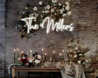 May include: A neon sign that reads "The Millers" is mounted on a brick wall, surrounded by floral arrangements. A wooden table is set with candles and floral centerpieces, creating a romantic ambiance for a wedding or event.