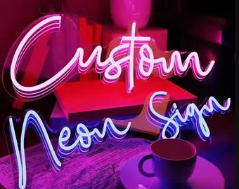 May include: A neon sign with the words "Custom Neon Sign" in white neon letters on a pink and purple background. The sign is sitting on a table with a cup of coffee and a book.