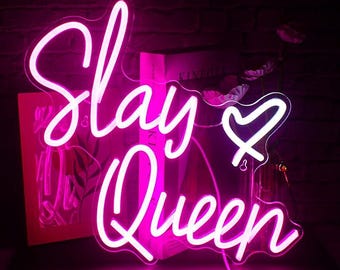 May include: A neon sign in a cursive font that reads "Slay Queen" with a heart symbol. The sign glows with a bright pink light. The sign is on a dark surface with a brick wall background.