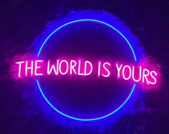 May include: A neon sign with the phrase "THE WORLD IS YOURS" in bright pink, encircled by a blue neon ring. The sign is set against a dark background with a fluffy, feather-like texture.