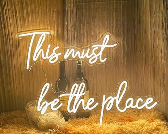 May include: A neon sign in white script reads "This must be the place" above two wine bottles. The sign is illuminated against a textured, golden-brown background. A fluffy, golden-brown surface is in the foreground.