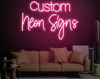 May include: A bright pink neon sign against a dark magenta wall. The sign reads "Custom Neon Signs" in a cursive font. Below the sign is a tan sofa with several pillows. A small side table and a cactus are also visible.