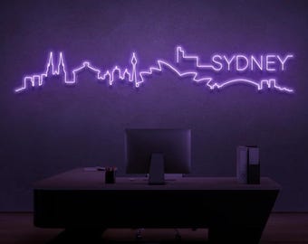 May include: A purple neon sign displaying the Sydney skyline. The sign is mounted on a dark purple wall above a desk with a computer monitor, office supplies, and a black desk. The word "SYDNEY" is written in neon.