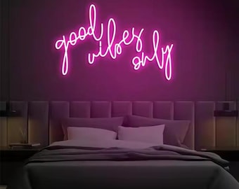 May include: A neon sign in cursive script that reads "good vibes only" in bright pink against a dark purple wall. The sign is above a bed with white pillows and a gray comforter. The room has a modern aesthetic.