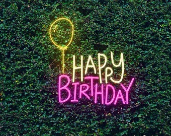 May include: A neon sign with the words "HAPPY BIRTHDAY" in pink and yellow, with a yellow balloon graphic, set against a backdrop of green foliage. The sign is illuminated, creating a festive and celebratory atmosphere.