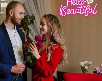 May include: A couple is smiling at each other, with the woman holding red roses. The man is wearing a blue suit jacket over a white shirt. The woman is wearing a red dress. A neon sign that says "Hello Beautiful" is on the wall.
