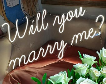 May include: A white neon sign that reads "Will you marry me?" is displayed. The sign is in a cursive font and is lit up. The sign is placed above a bouquet of white roses with green leaves. The background is a soft, neutral color.