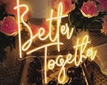 May include: A neon sign with the words "Better Together" in a cursive font. The sign is illuminated with a warm yellow glow. The background features pink flowers and a book.