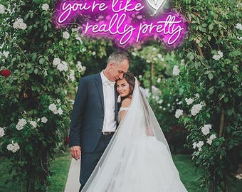 May include: A couple stands beneath a floral archway, the bride in a white wedding dress and veil, the groom in a navy suit. Above them, a neon sign reads "you're like really pretty" in pink and white, surrounded by white flowers.