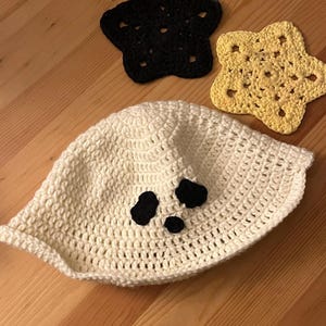 May include: A white crocheted bucket hat featuring two black oval eyes and a small black circular mouth. Accompanying the hat are two star-shaped crocheted coasters, one in solid black and one in pale yellow, with intricate openwork patterns.