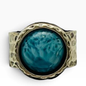 May include: A ring featuring a large, round, teal-colored stone with a marbled appearance, set within a wide, hammered-texture metal band that has a darkened, antique-style finish around the edges and the setting.