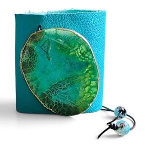 May include: A wide turquoise leather cuff bracelet featuring a large, oval-shaped green agate slice with a gold-toned metallic edge. The bracelet includes two thin black cords extending from the side, each finished with a small, translucent blue bead.