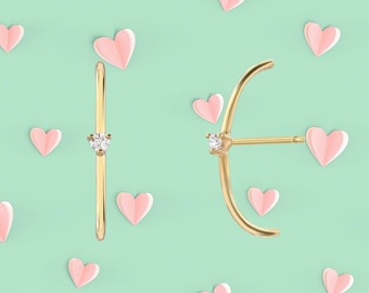Pode incluir: Dois brincos dourados com um pequeno detalhe de diamante são exibidos em um fundo verde claro com recortes de coração rosa. Um brinco é uma barra reta, e o outro tem um design curvo.
