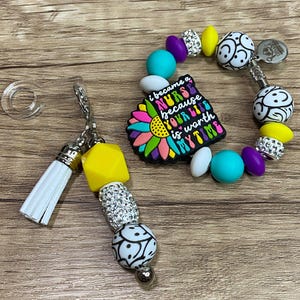 May include: A beaded bracelet and keychain set with a nurse theme. The bracelet features a charm with the text "I became a nurse because your butt is worth my time." The keychain has a white tassel and yellow and silver beads.