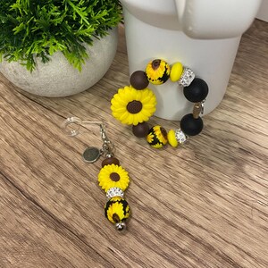 May include: A sunflower-themed bracelet and a matching accessory. The bracelet features yellow sunflower beads, black beads, and crystal accents. The accessory has a sunflower charm, crystal accents, and a small metal charm.