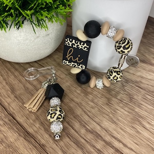 May include: A beaded keychain and bracelet set. The keychain features a tan tassel, black geometric bead, and leopard print bead. The bracelet has black, tan, and leopard print beads, with a black rectangle that says "hi".