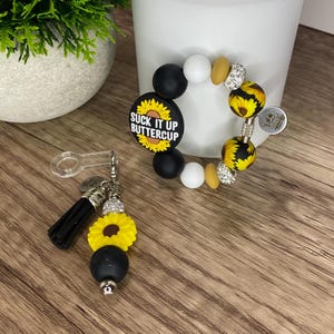 May include: A beaded bracelet and a keychain with a sunflower theme. The bracelet features black, white, yellow, and rhinestone beads. The keychain has a black tassel, a yellow sunflower, and a black bead. The bracelet has a sunflower-shaped charm with the text "Suck It Up Buttercup".