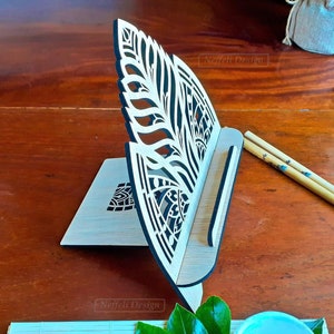 Puede incluir: Un soporte de teléfono de madera con un diseño de mandala. El soporte está hecho de madera clara con detalles negros. El soporte tiene un grosor de 3 mm o 1/8 de pulgada. El soporte está sobre una superficie de madera.