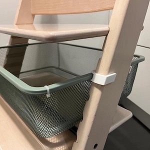 May include: A white wooden high chair with a green wire mesh basket attached to the bottom. The basket is designed to hold toys or other items.
