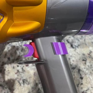 May include: Close-up of a Dyson vacuum cleaner. The vacuum features a gold and gray body with purple and red accents. A purple clip is attached to the gray handle. The image highlights the design and color scheme of the appliance.