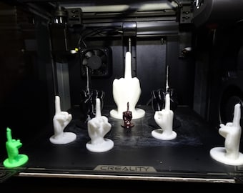 May include: A 3D printed model of a middle finger, printed in white, black, and green plastic. The model is on a black surface with the word "CREALITY" printed on it.