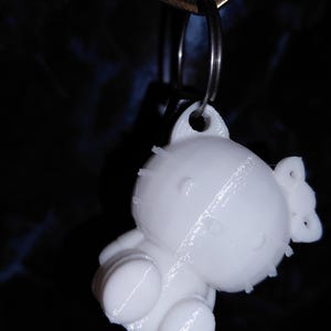 Peut inclure: Un porte-clés blanc imprimé en 3D représentant un personnage de dessin animé. Le porte-clés est attaché à une clé en métal. Le design comprend un nœud et des formes arrondies. La clé porte le mot "GUARD".