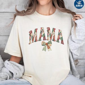 Peut inclure: T-shirt couleur ivoire avec le mot "MAMA" dans un motif patchwork, avec des motifs rouges, verts et blancs. Un motif de nœud avec des motifs similaires se trouve en dessous. Le t-shirt est de style décontracté et confortable.