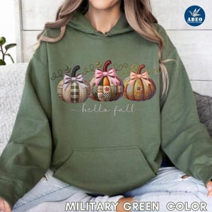 Peut inclure: Sweat à capuche vert militaire avec un motif de trois citrouilles décoratives avec des nœuds et le texte "hello fall". Les citrouilles ont des motifs patchwork de différentes couleurs. Le sweat à capuche a une poche avant.