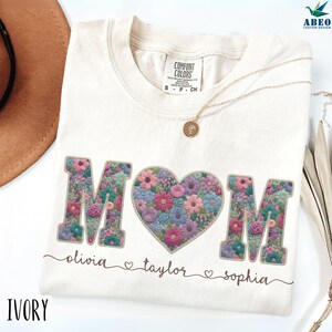 Peut inclure: T-shirt ivoire avec le mot "MOM" en imprimé floral, un motif floral en forme de cœur et les noms "olivia", "taylor" et "sophia" en dessous. Un collier en or avec un pendentif est sur le t-shirt.