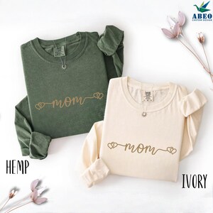 Peut inclure: Deux t-shirts à manches longues, un vert chanvre et un ivoire, avec le mot "maman" en écriture cursive, flanqué de cœurs. Les chemises ont un col rond et sont présentées sur une surface blanche.
