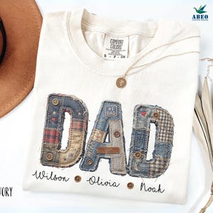 Peut inclure: T-shirt ivoire avec le mot "DAD" en patchwork. Les lettres sont faites de différents tissus, dont du denim et du plaid. Les noms "Wilson", "Olivia" et "Noah" sont imprimés en dessous. Un collier en or est drapé sur le t-shirt.