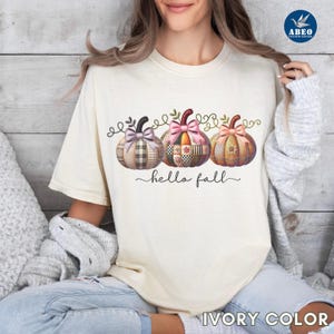 Peut inclure: T-shirt couleur ivoire avec trois citrouilles décoratives et l'inscription "hello fall". Les citrouilles présentent des motifs patchwork dans des tons de rose, marron et orange, avec des nœuds. Le t-shirt est de style décontracté.