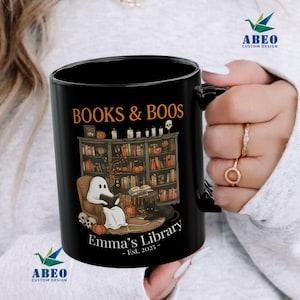 Peut inclure: Mug en céramique noire avec un motif sur le thème d'Halloween. Le mug affiche le texte "BOOKS & BOOS" au-dessus d'une scène de bibliothèque avec un fantôme lisant, un chat noir et des citrouilles. Le texte "Emma's Library - Est. 2025-" est également présent.