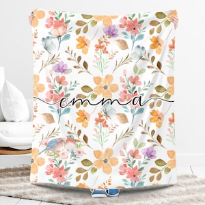 May include: A white blanket with a floral design in orange, pink, and purple. The blanket has the name "emma" written on it in a cursive font. Two blue birds are also visible.
