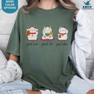 May include: Green t-shirt with three cartoon lucky cats holding gold coins and wearing red collars. The text "good luck - good life - good vibes" is printed below. The shirt has a loose fit and is made of a soft material.