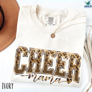 Peut inclure: T-shirt ivoire avec l'inscription "CHEER mama" dans un motif marron et léopard. Un collier doré avec un pendentif est posé sur le t-shirt. Le t-shirt est de la marque Comfort Colors.