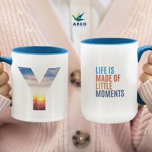Peut inclure: Deux tasses en céramique blanche avec des bords bleus. Une tasse présente une image colorée d'un coucher de soleil à l'intérieur de la lettre Y. L'autre tasse porte le texte "Life is made of little moments" en texte coloré.
