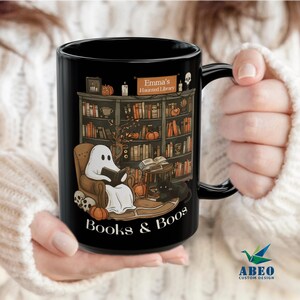 Peut inclure: Mug en céramique noire avec une scène de bibliothèque effrayante, avec un fantôme lisant un livre, des citrouilles et un chat noir. Le mug porte l'inscription "Books & Boos" et "Emma's Haunted Library".