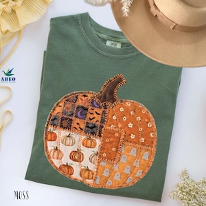 Peut inclure: Un t-shirt à col rond vert mousse arborant un grand motif central de citrouille composé de divers empiècements de style tissu. Le design de la citrouille inclut des motifs de petites citrouilles, fantômes, chauves-souris, lunes et toiles.