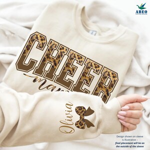Peut inclure: Sweat-shirt crème avec le mot "CHEER" en lettres marron, avec un motif léopard. Le mot "mama" est en dessous en écriture cursive. La manche présente le nom "Olivia" et un nœud léopard.