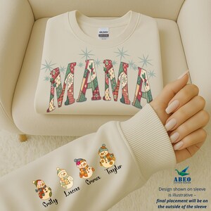Peut inclure: Sweat-shirt crème avec le mot "MAMA" dans un motif patchwork. La manche présente quatre illustrations de bonhommes de neige avec les noms : Emily, Lucas, Emma et Taylor. Le design comprend des accents de flocons de neige, parfait pour les fêtes.