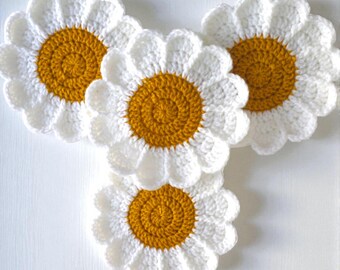 May include: Four handmade crochet daisy coasters. Each coaster features a white scalloped edge and a golden yellow center. The coasters are arranged on a white surface, showcasing their floral design and texture.