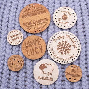 May include: A collection of eight round cork labels with various designs and text. The labels feature designs such as mountains, a paw print, a snowflake, a sheep, and text such as "Mountain Wear", "Love Lucy", "Happy Holidays", "100% Merino Wool", and "By Maggie".