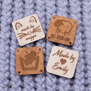 May include: Four square cork fabric labels with different designs. One label has a cat with the text "made by moggie". Another label has a sheep with the text "100% Merino Wool". A third label has a hand washing symbol. The final label has the text "Made by Emily" with a heart.