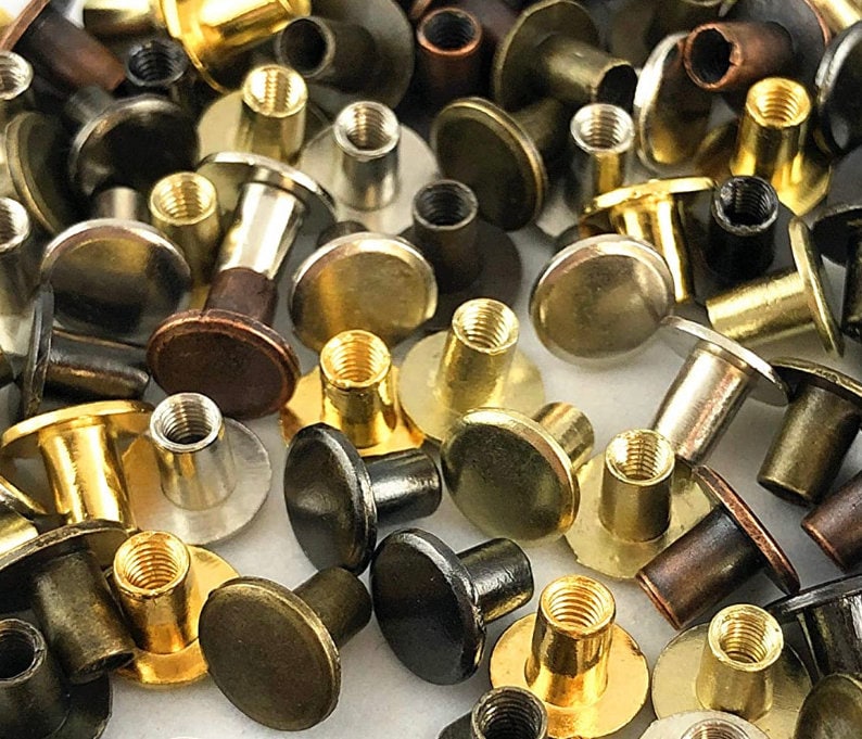 Chicago screws are shown in various colors offered, strewn across a white table. Colors include Silver, Gold, Bronze, Copper, Gray metal