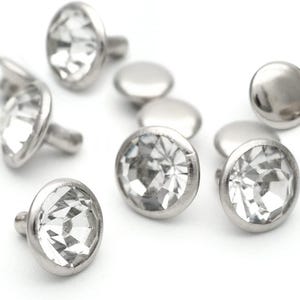 May include: A close-up of a group of silver-toned metal studs with clear crystal accents. The studs have a flat, round base and a small, pointed post.