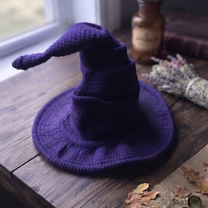 May include: A deep purple crocheted hat with a wide, circular brim and a tall, pointed crown that curves downward at the tip. The hat features a textured, yarn-based construction with visible stitches throughout the entire surface of the object.